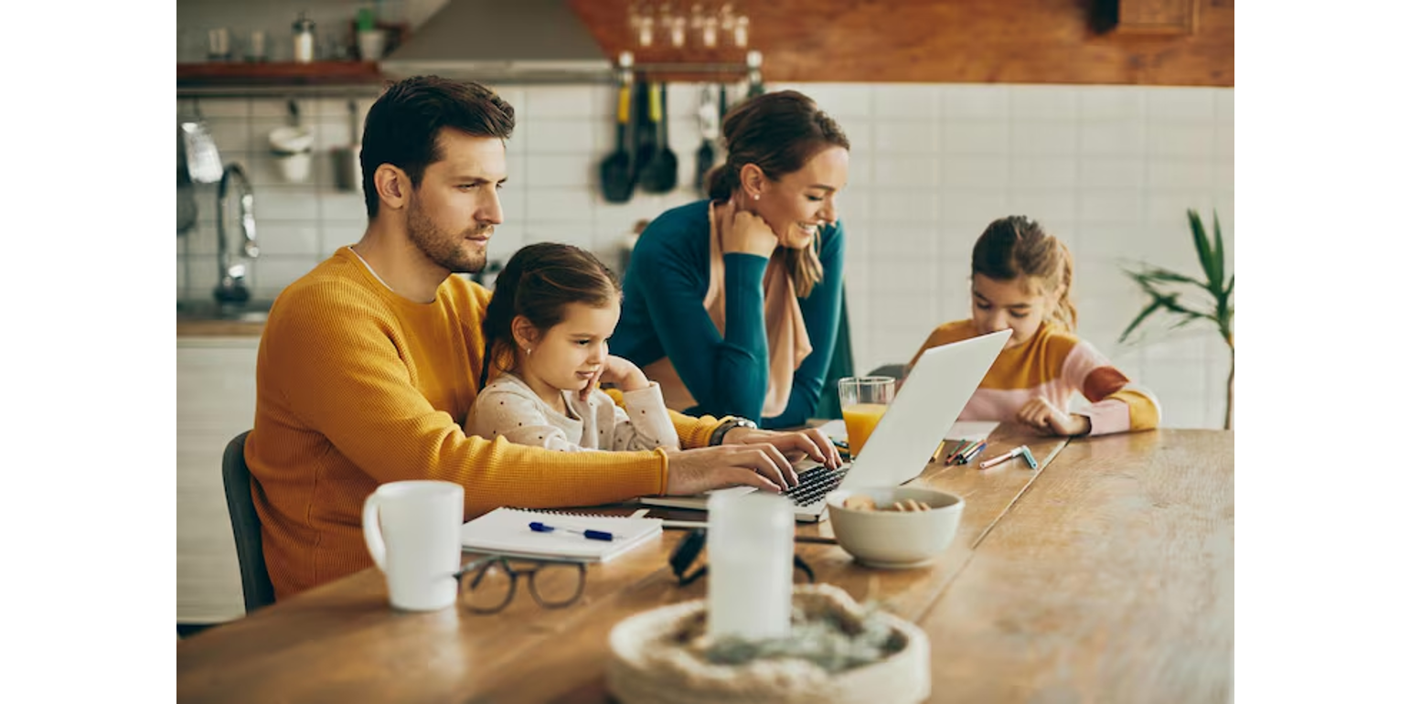 Family of four at a kitchen table, father working on a laptop with daughter on his lap, mother smiling nearby, and another daughter coloring with markers.