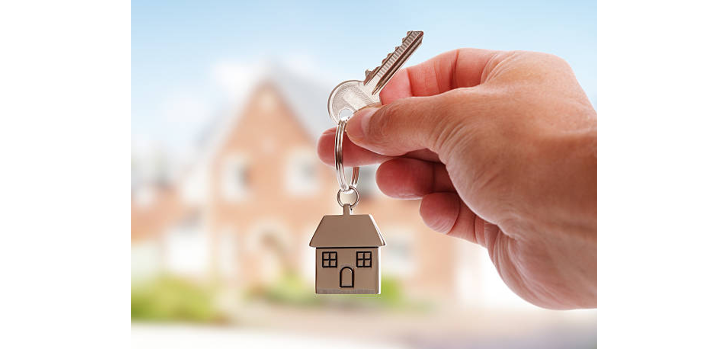 Hand holding house key with home-shaped keychain in front of a blurred brick house, symbolizing new home ownership.