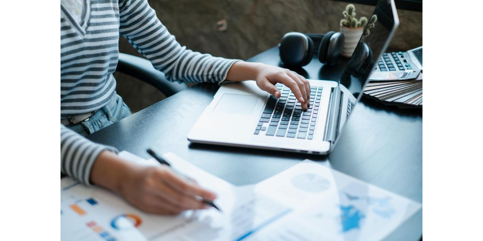 Person in a striped shirt working on a laptop while taking notes on printed charts at a desk with headphones.