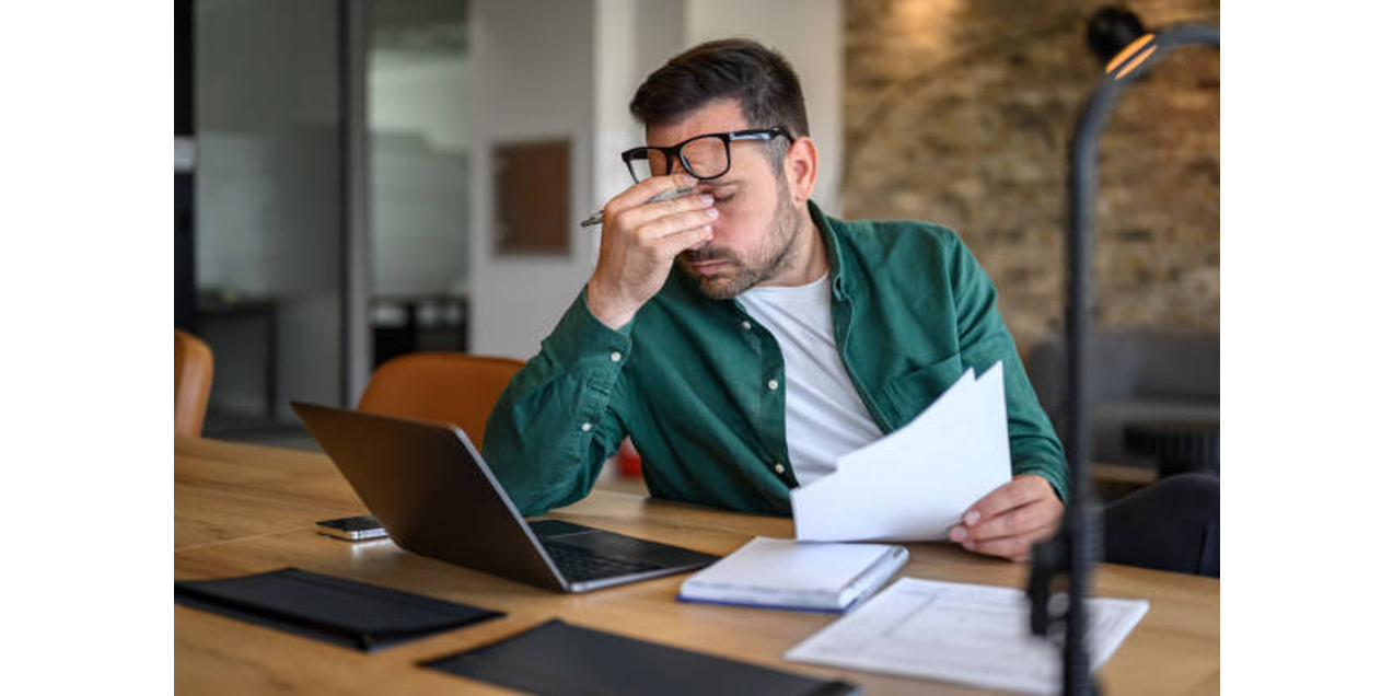 Stressed man in green shirt rubbing his eyes while holding documents at desk with laptop, appearing frustrated or exhausted from work.