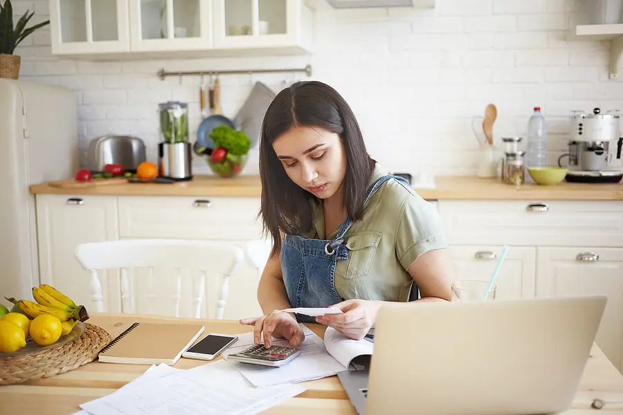 Young woman using a calculator while reviewing bills and paperwork at her kitchen table with a laptop, managing household finances.