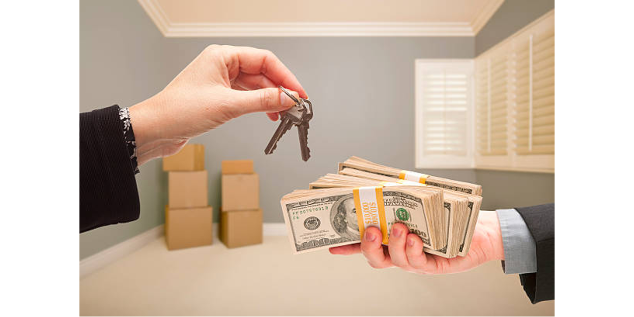 Hand exchanging house keys for a stack of cash in an empty room with moving boxes in the background.