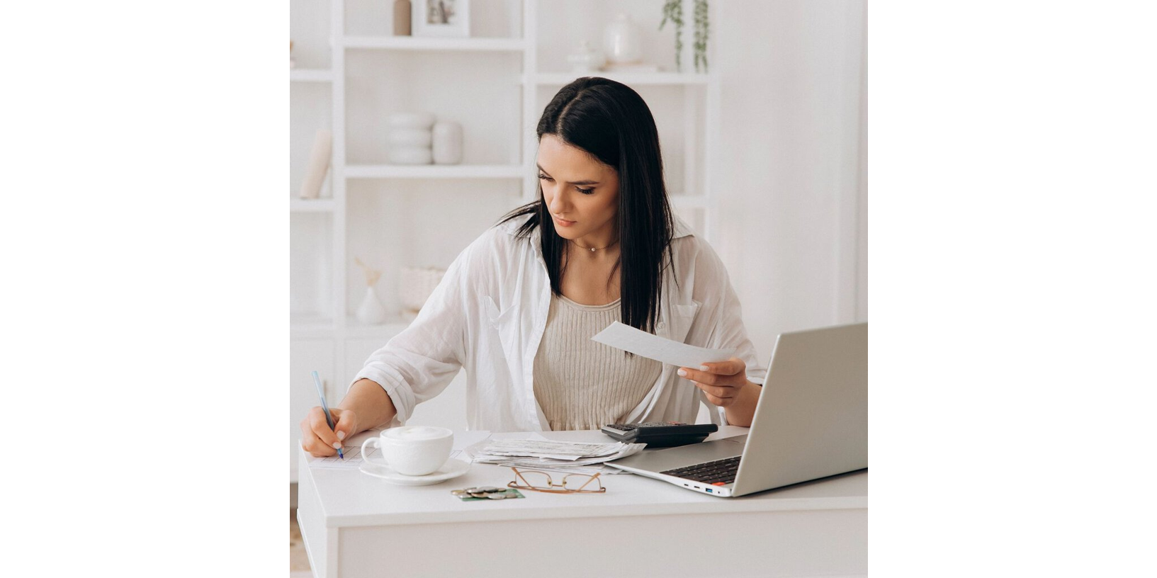 Woman reviewing bills and taking notes at a white desk with a laptop, calculator, and coffee cup.