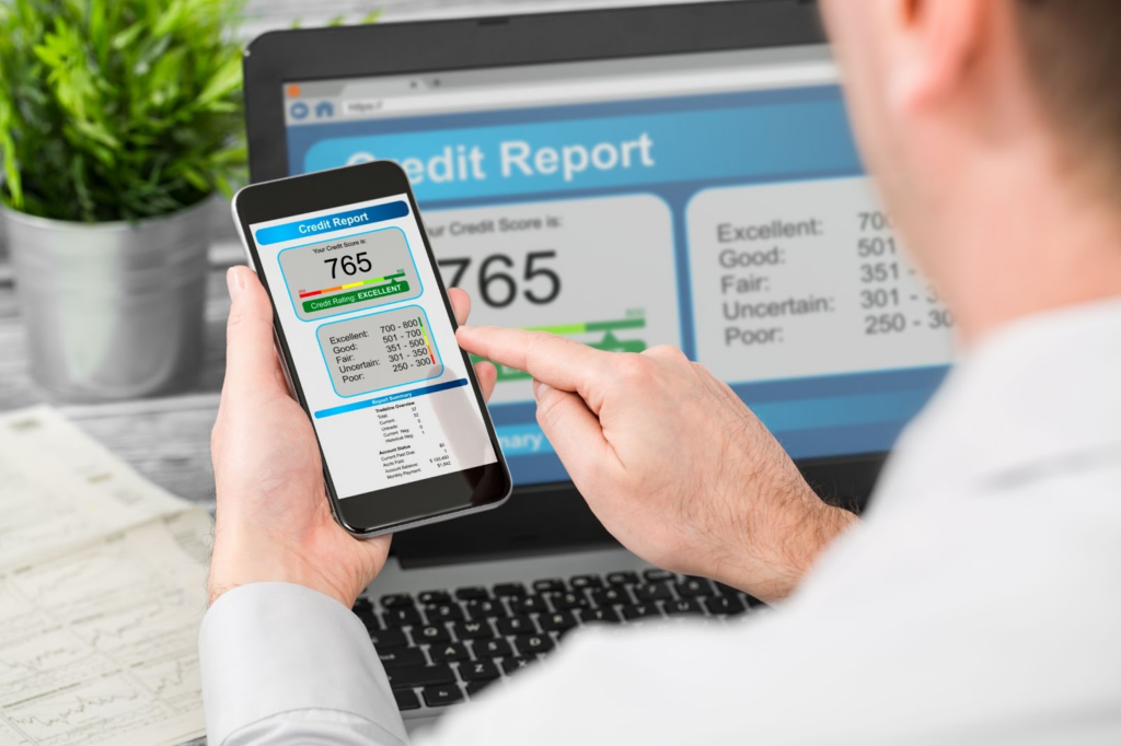 Person checking credit report with 765 score on smartphone while same information displays on the laptop screen in a background.