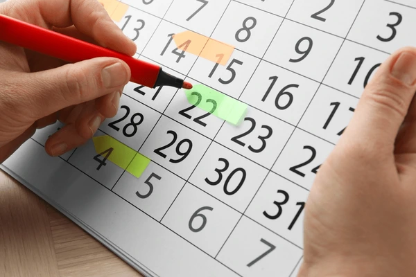 Hands marking a date on a paper calendar with a red marker, colorful sticky tabs highlighting important dates.