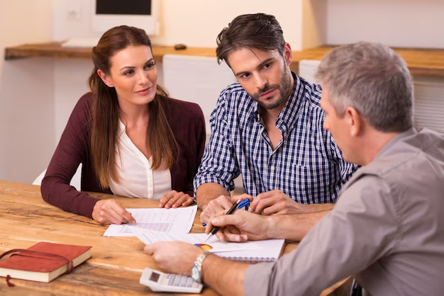 Young couple meeting with advisor at wooden table, reviewing and signing paperwork together with documents and calculator visible.