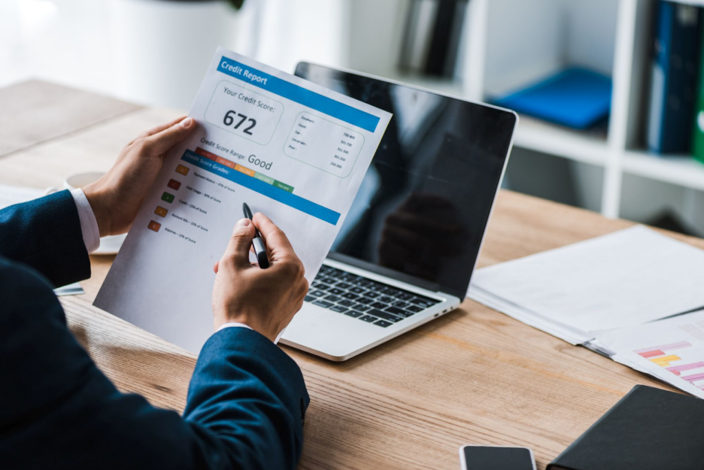 Business professional in a suit reviewing printed credit report showing score of 672 rated as "Good" at the desk with a laptop.