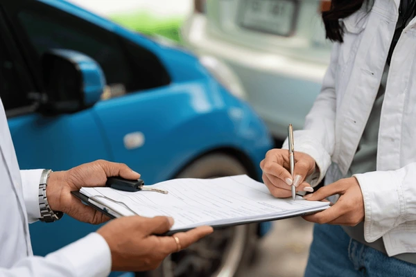 Person signing paperwork on clipboard held by salesperson with car keys.