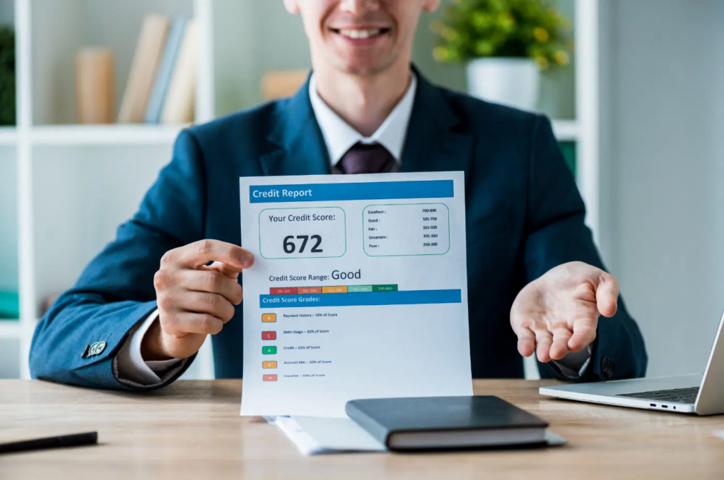 Smiling businessman holding up a credit report showing a score of 672 with "Good" rating while gesturing at his desk with a laptop.