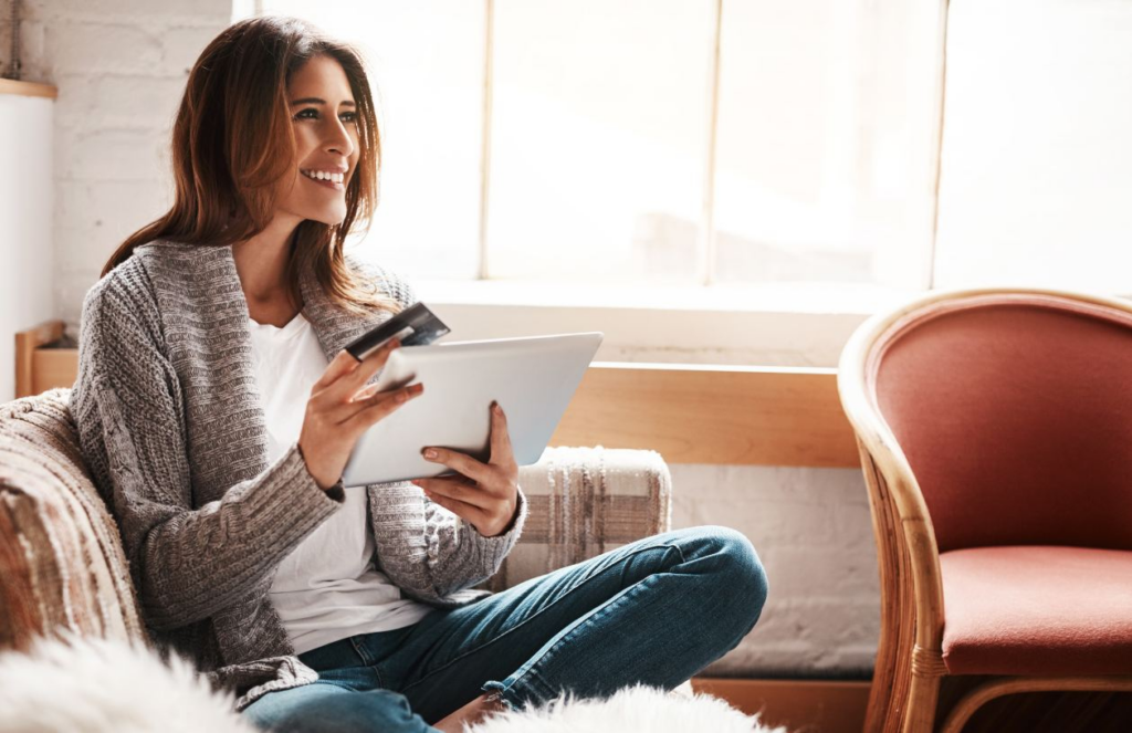 Woman in gray cardigan smiling while holding a tablet and a credit card, sitting on a couch in a sunny living room.
