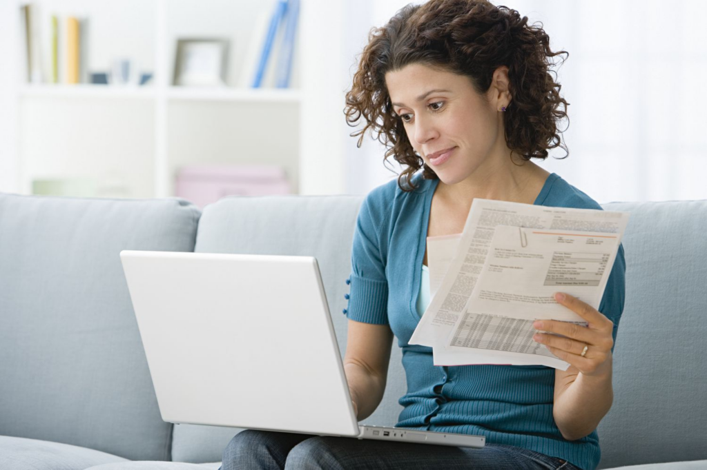 Woman with curly hair holding bills while using a laptop on a gray couch at home.