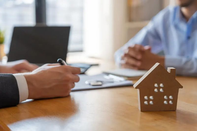 Wooden house model on a desk during a real estate meeting with two people reviewing documents in the background.