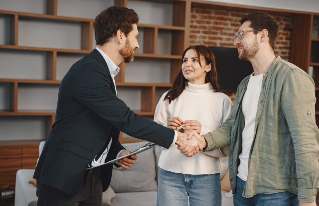 Real estate agent handing keys to a smiling couple while shaking hands in a modern living room.